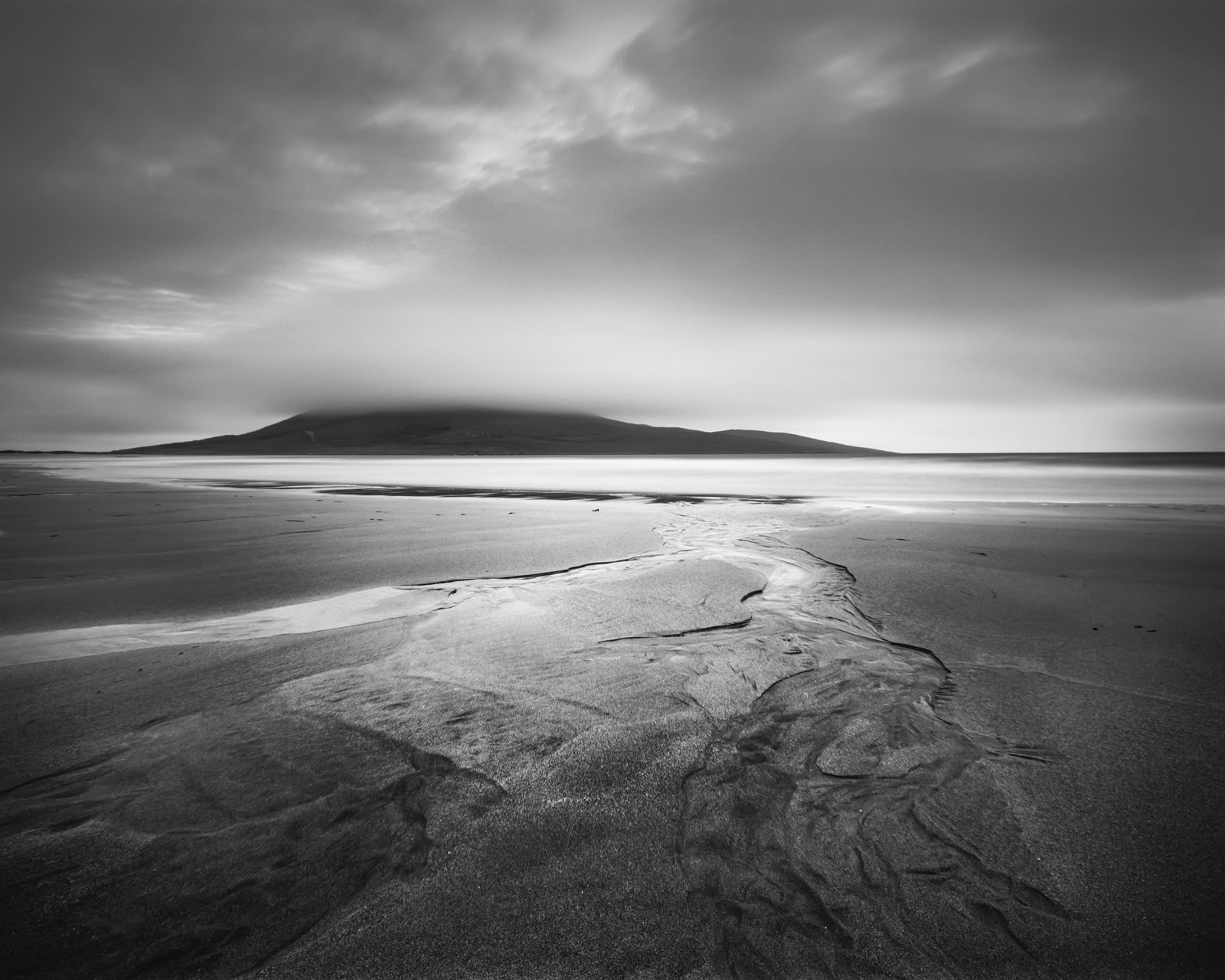 A monochrome landscape featuring a serene beach with smooth sand intersected by a meandering stream. In the backdrop, a gentle hill shrouded in mist rises under a cloudy sky. The scene conveys tranquillity and the vastness of nature.