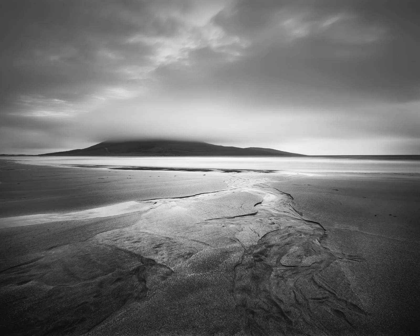 Monochrome coastal landscape with tidal sand patterns, shallow water channels, and a dark volcanic island beneath overcast sky.
