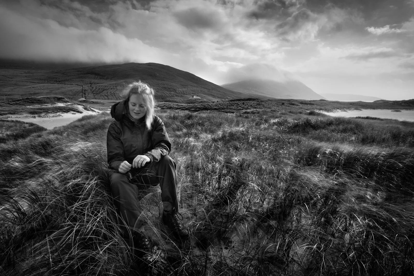 Woman in black jacket crouching among moorland grasses with mountains and dramatic sky behind.
