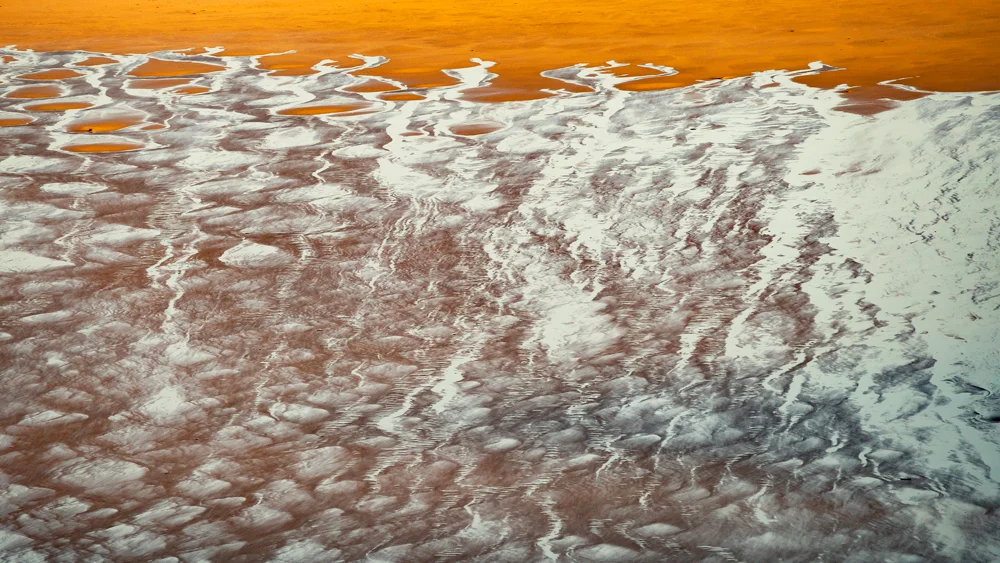 Orange salt lake with white mineral deposits and intricate patterns forming natural ridges and channels along the shoreline.