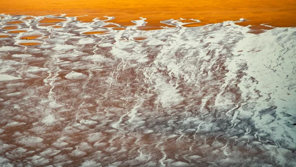 Orange salt lake with white mineral deposits and intricate patterns forming natural ridges and channels along the shoreline.