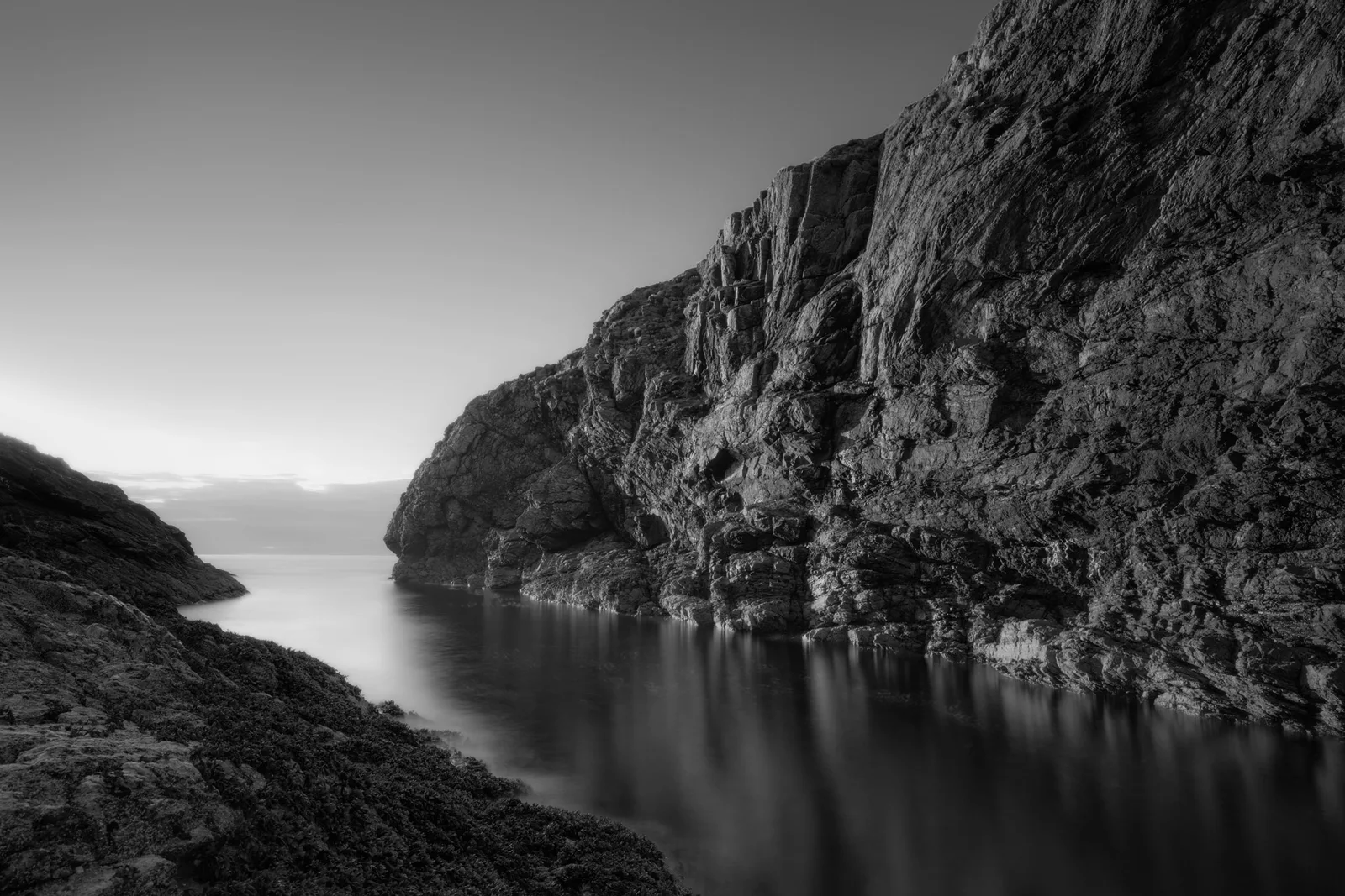 Black and white seascape of sheer limestone cliffs rising from still water, with distant headland under grey overcast sky.