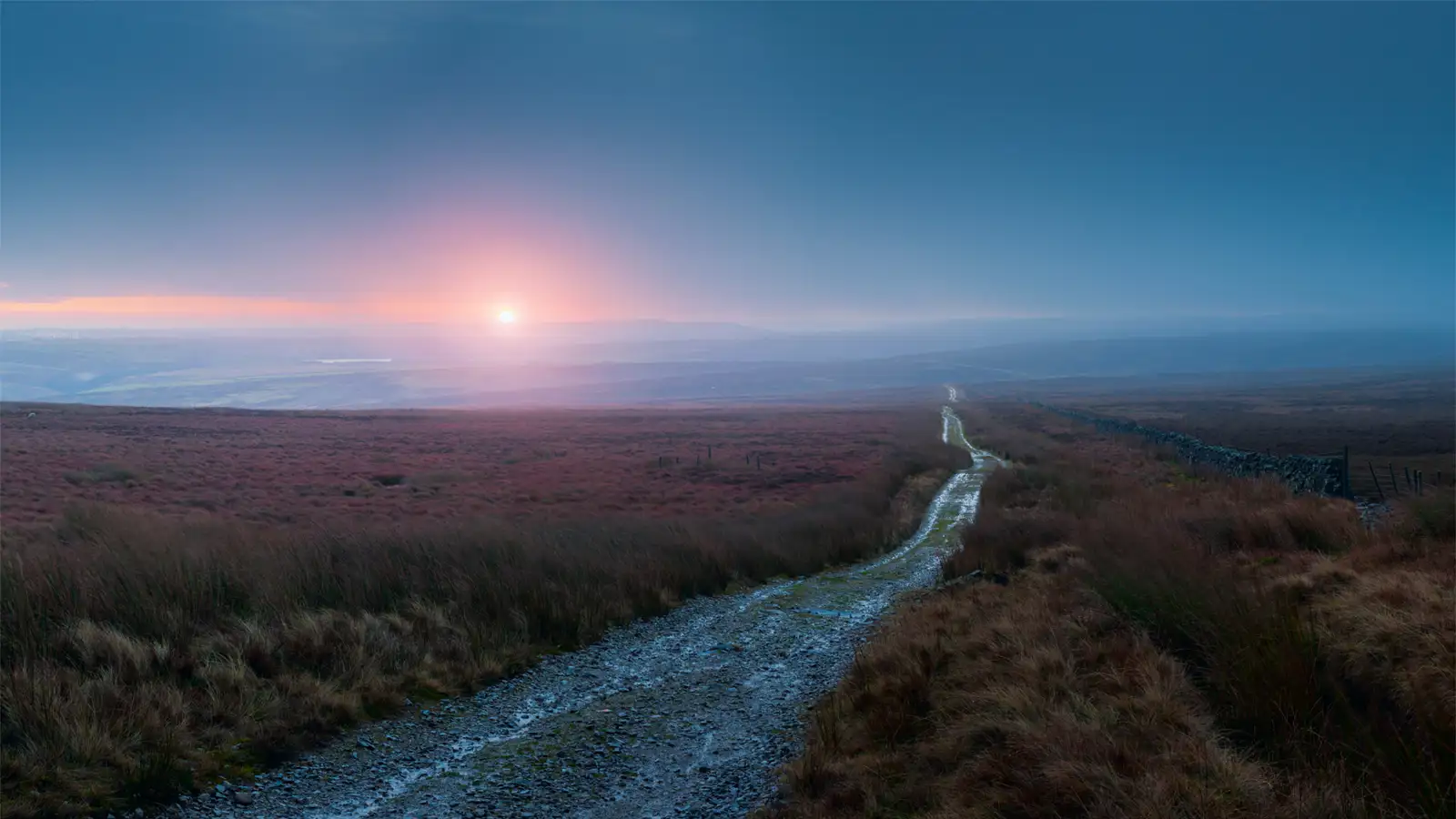 Pathway leading through an expansive moorland at sunrise, with a stone wall running parallel to the path on the right. The sky transitions from a deep blue to a warm pink near the horizon, where the sun is partially obscured by haze. Vegetation on the moor is sparse, dominated by grasses and low shrubs.