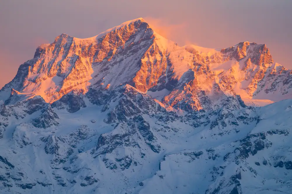Snow-covered mountain peaks bathed in warm, golden sunlight during sunset. The light creates a striking contrast with the deep blue shadows in the crevices and valleys, highlighting the rugged texture and grandeur of the landscape.