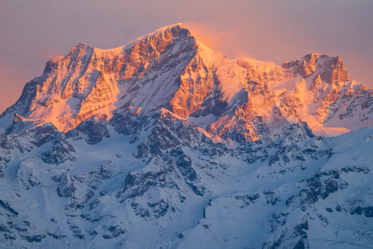 Snow-covered mountain peaks bathed in warm, golden sunlight during sunset. The light creates a striking contrast with the deep blue shadows in the crevices and valleys, highlighting the rugged texture and grandeur of the landscape.