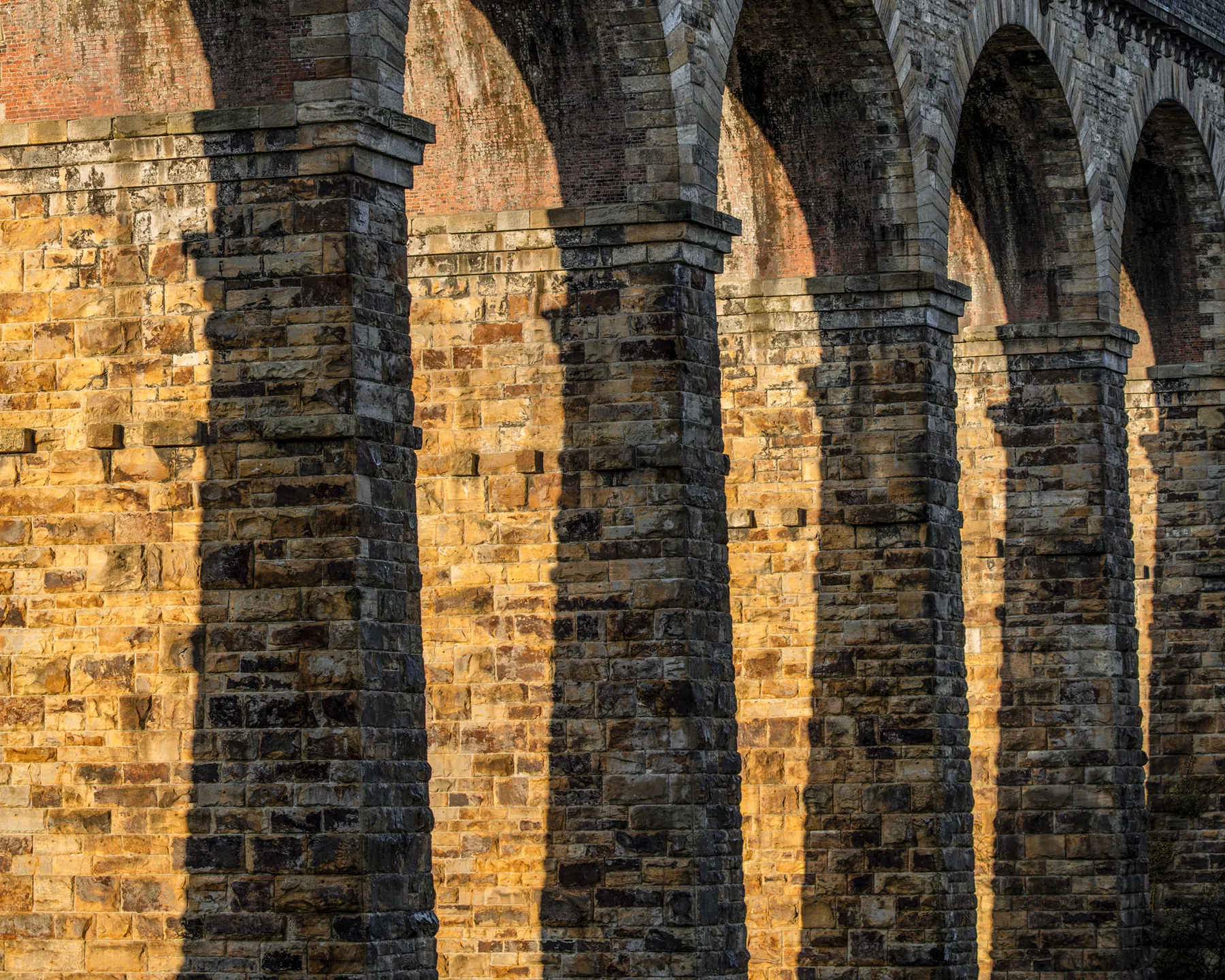 Stone railway viaduct with multiple arched spans and pillars, lit by warm golden sunlight casting shadows.