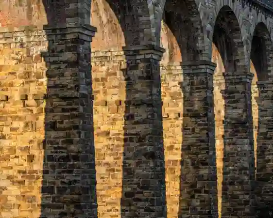 The Crimple Valley Viaduct's stone structure bathed in sunlight. Arches rise from sturdy pillars, their shadows striping the textured walls. The golden hue of the stone contrasts with the soft arch shadows, evoking a sense of historic grandeur.