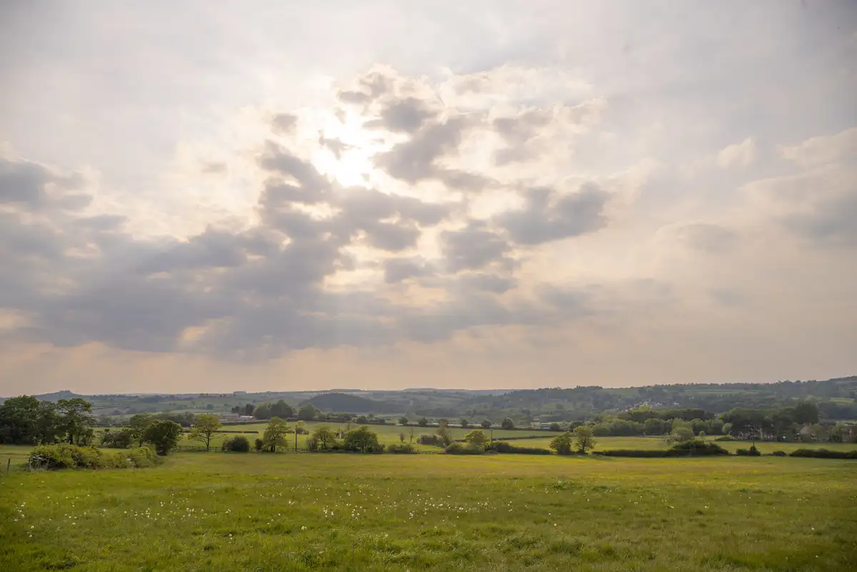 Rolling countryside landscape with green fields stretching into the distance. Sunlight filters through scattered clouds, casting soft rays on the land below. Sparse trees and hedgerows border the fields, with hills visible on the horizon under a partly cloudy sky.