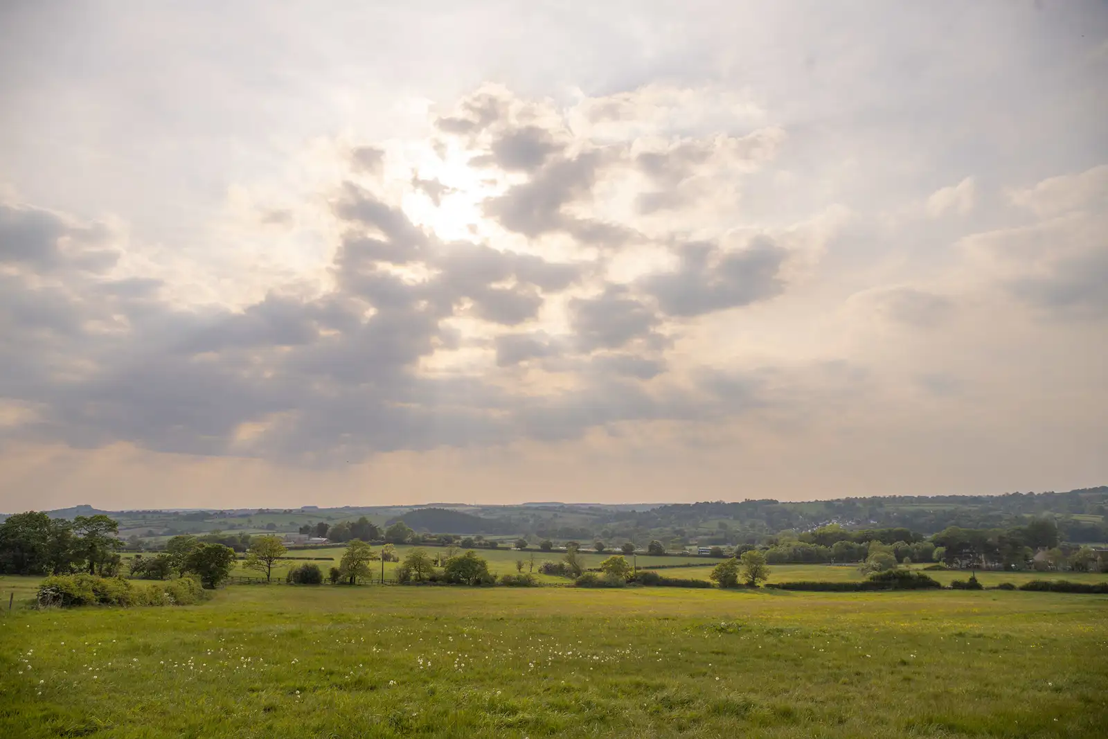 Rolling countryside landscape with green fields stretching into the distance. Sunlight filters through scattered clouds, casting soft rays on the land below. Sparse trees and hedgerows border the fields, with hills visible on the horizon under a partly cloudy sky.
