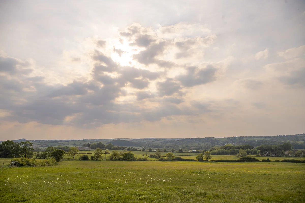 Rolling countryside meadow with wildflowers beneath a broken sky as sunlight breaks through cloud at dusk