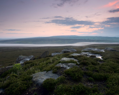 Moorland Landscapes: Mist hovers above a moorland valley in North Yorkshire, which blends into a soft colour palette of dusky pinks and blues in the sky. Foreground rocks and grasses provide texture against the sweeping view of this serene landscape. a close up of a hillside next to a body of water