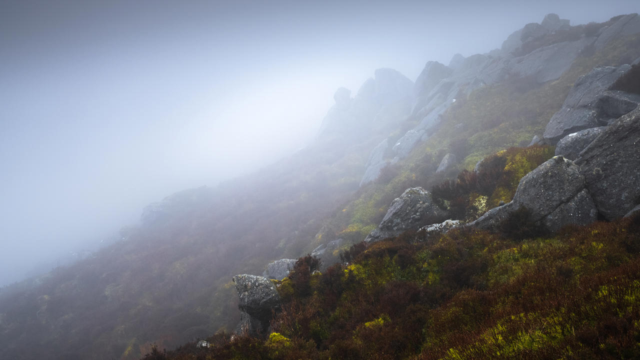 Foggy hillside with large grey boulders and patches of green and brown vegetation. The mist obscures the upper part of the hill, creating a mysterious atmosphere.