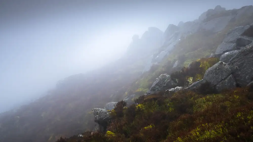 Foggy hillside with large grey boulders and patches of green and brown vegetation. The mist obscures the upper part of the hill, creating a mysterious atmosphere.
