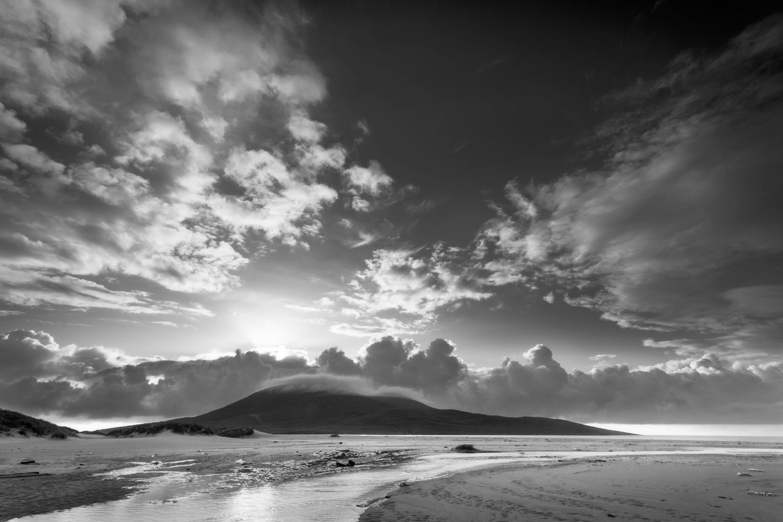 Black and white landscape of a coastal scene featuring a wide expanse of sandy beach in the foreground with gentle tidal pools. A low, grassy dune rises on the left, leading to a hill in the background. Dramatic cloud formations fill the sky, partially obscuring the sun and creating a striking contrast.