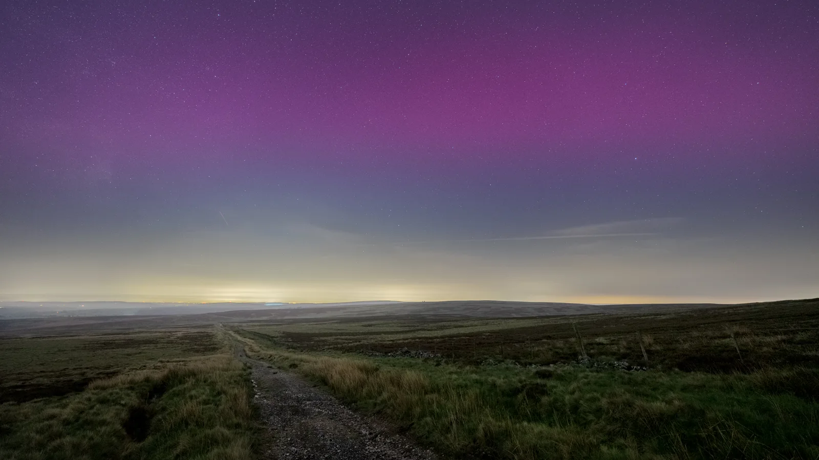 Moorland landscape at twilight with a dirt track, purple aurora sky above rolling green hills and distant horizon.