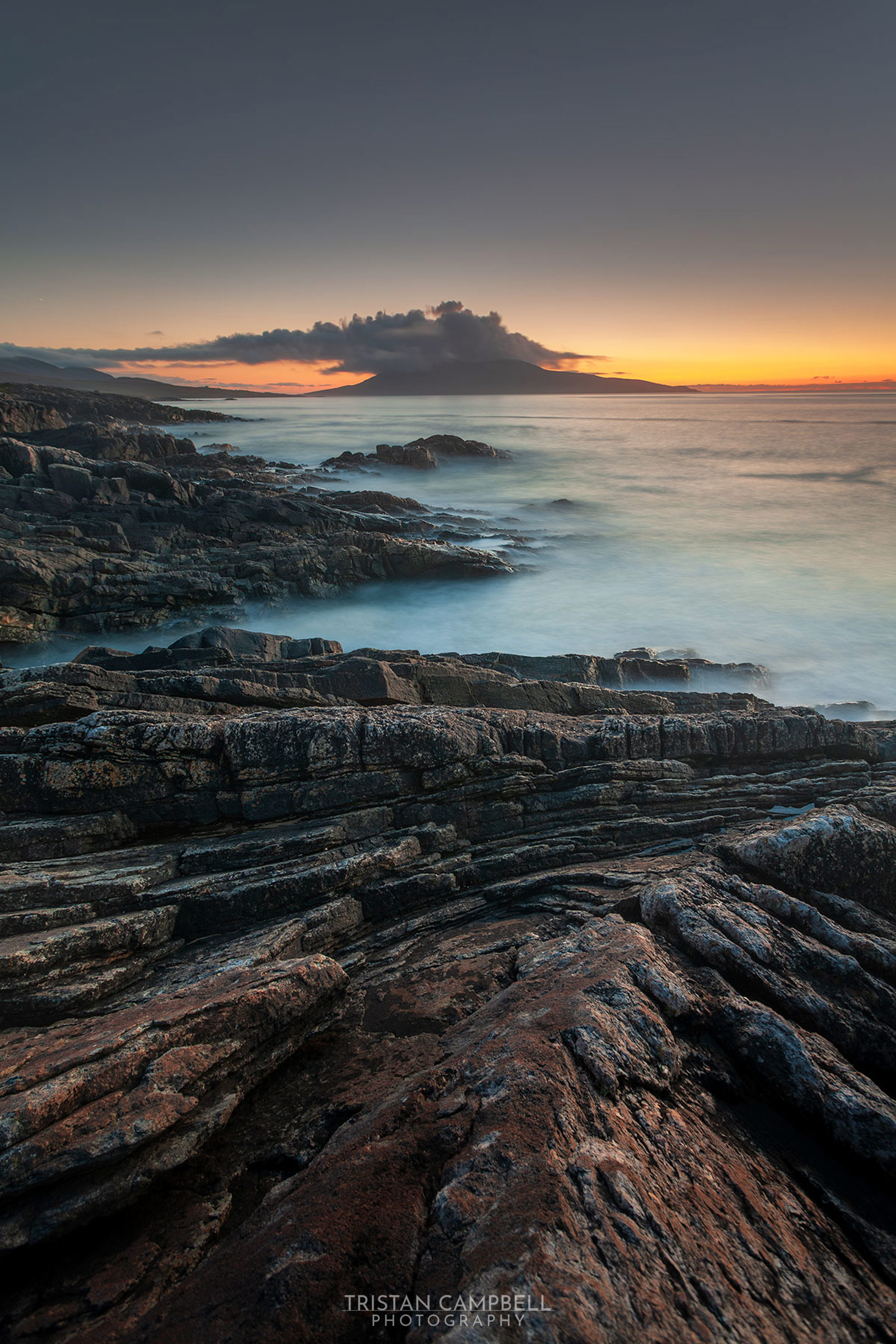 Rocky coastline with layered, textured stones leading to a calm sea. In the distance, a silhouetted island sits under a sky transitioning from deep blue to a warm orange at the horizon, as clouds hover above during sunset.