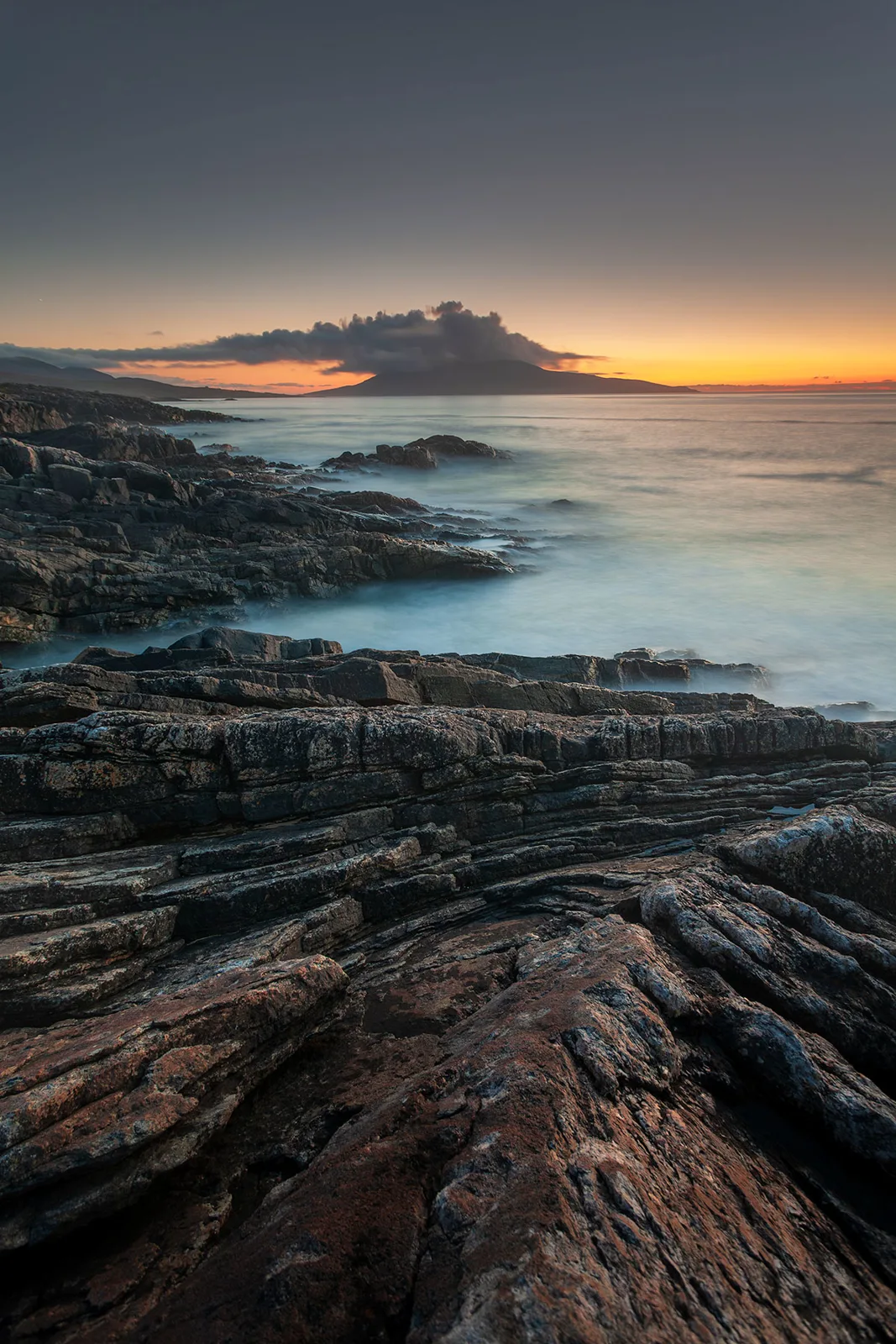 Rocky coastal foreground at sunset with calm sea and dramatic cloud formation on horizon beneath golden and blue sky.