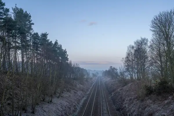 Railway tracks stretch into the distance, flanked by bare trees on both sides. The scene is set in early morning with a light frost covering the ground. The sky is clear with a hint of pink on the horizon, suggesting sunrise.