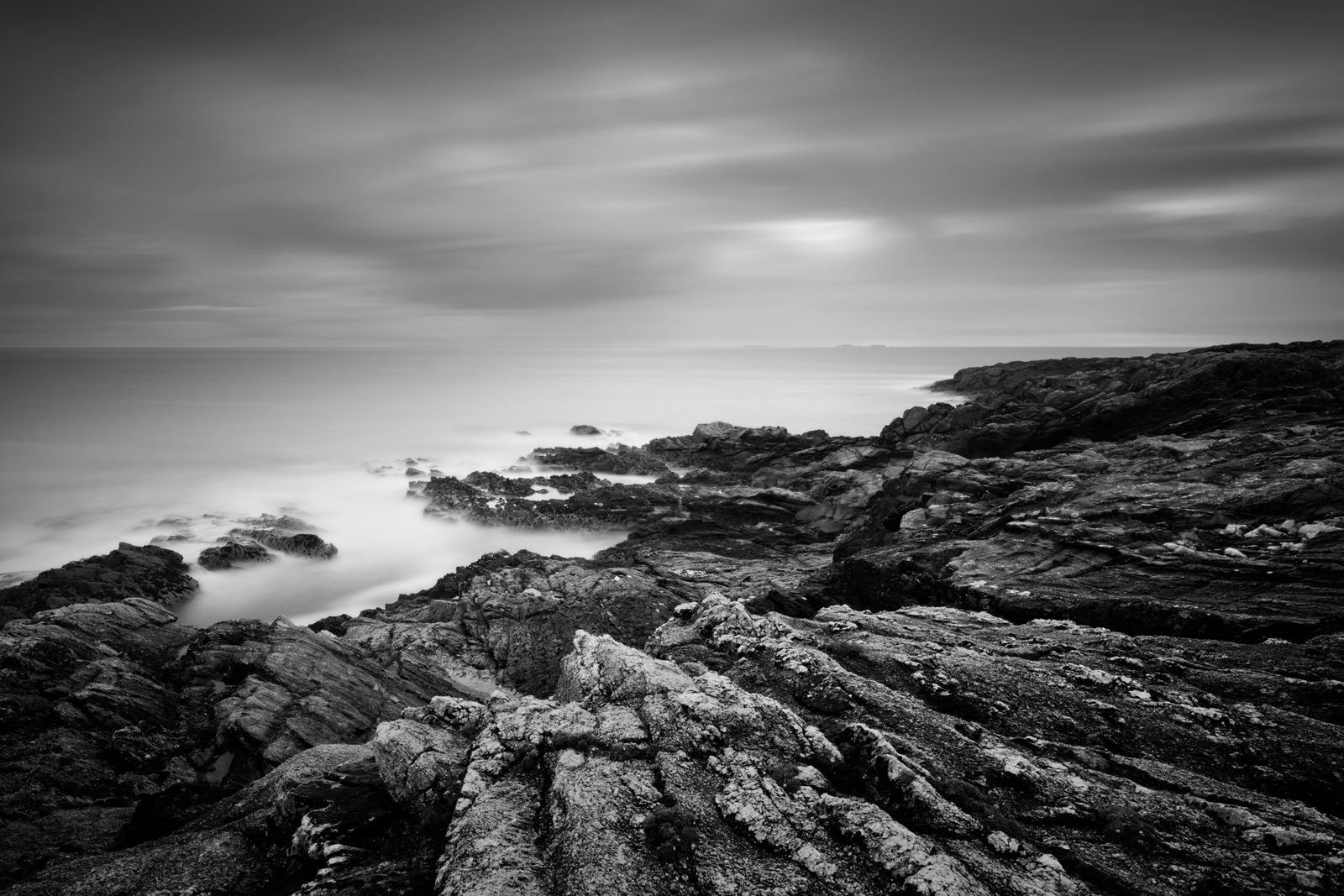 A monochrome seascape presents textured rocks in the foreground, leading into a serene ocean. Mist veils the water, and a tranquil sky with wispy clouds stretches above. A hint of landform is visible on the horizon.