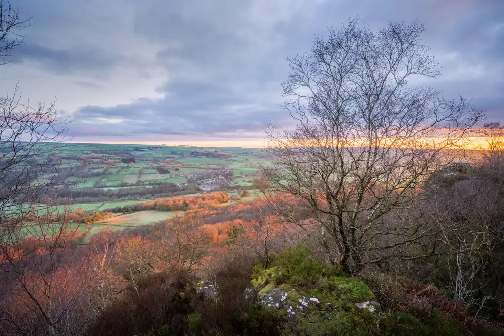 Rolling countryside landscape at sunset, with a foreground of bare trees and rocky ground. The sky is cloudy, with pink and orange hues along the horizon. Farmland fields stretch across the valley, dotted with patches of woodland.