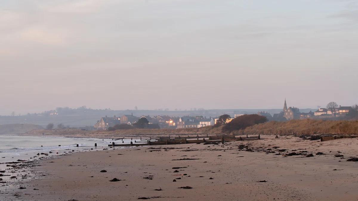 Wooden groynes on a sandy beach at dusk with a coastal village and church spire glowing softly in the distance
