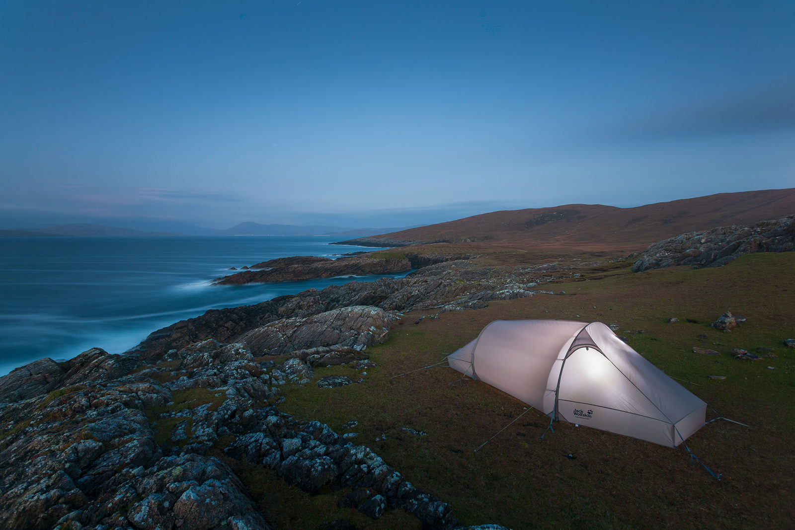 A solitary tent is pitched on a rugged, grassy cliffside overlooking the sea. The smooth rocks and gentle waves create a tranquil scene under the soft blue hues of twilight. The shoreline stretches into the distance with rolling hills visible under the expansive evening sky.