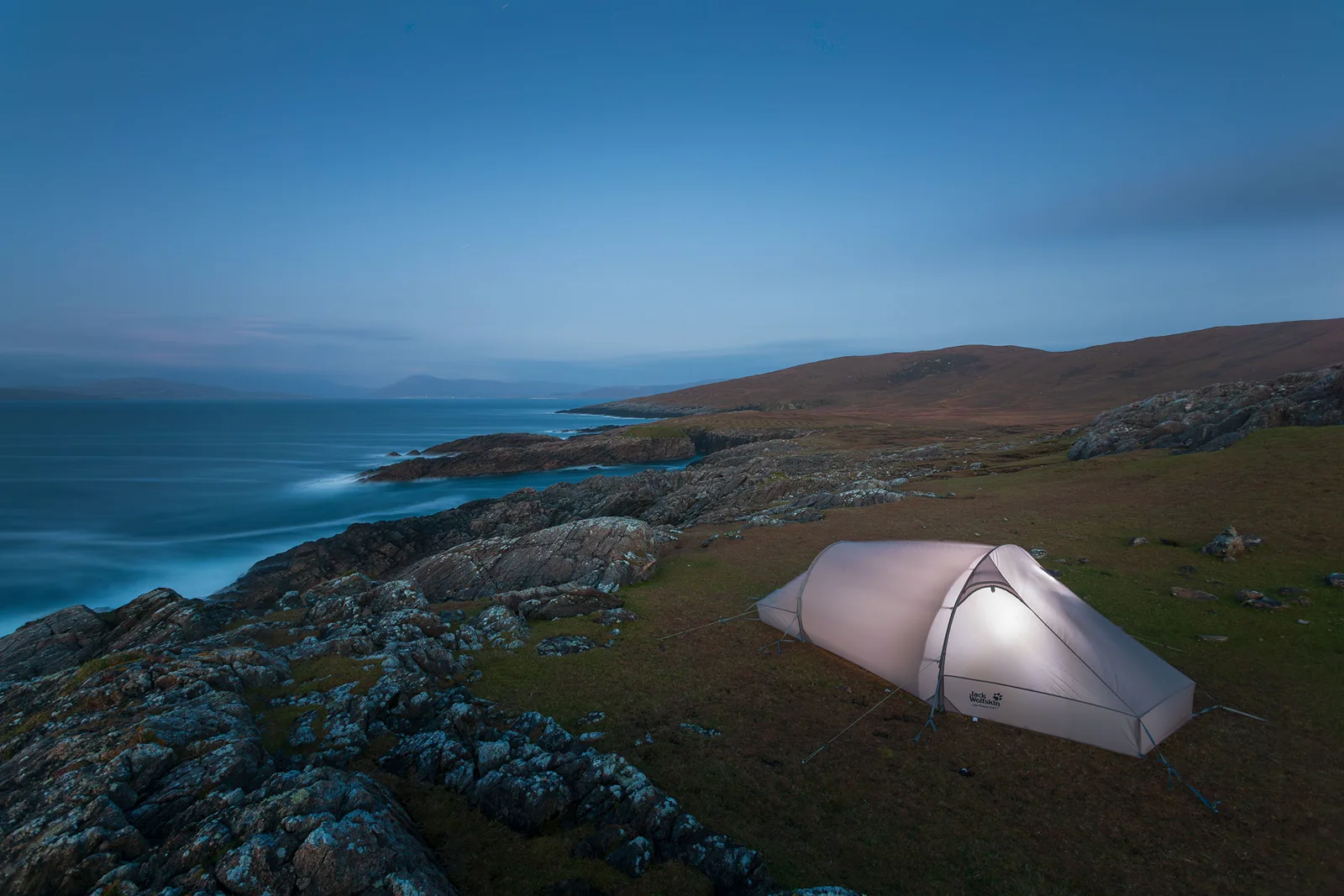 White tent pitched on moorland overlooking rocky coastline and turquoise sea at dusk.