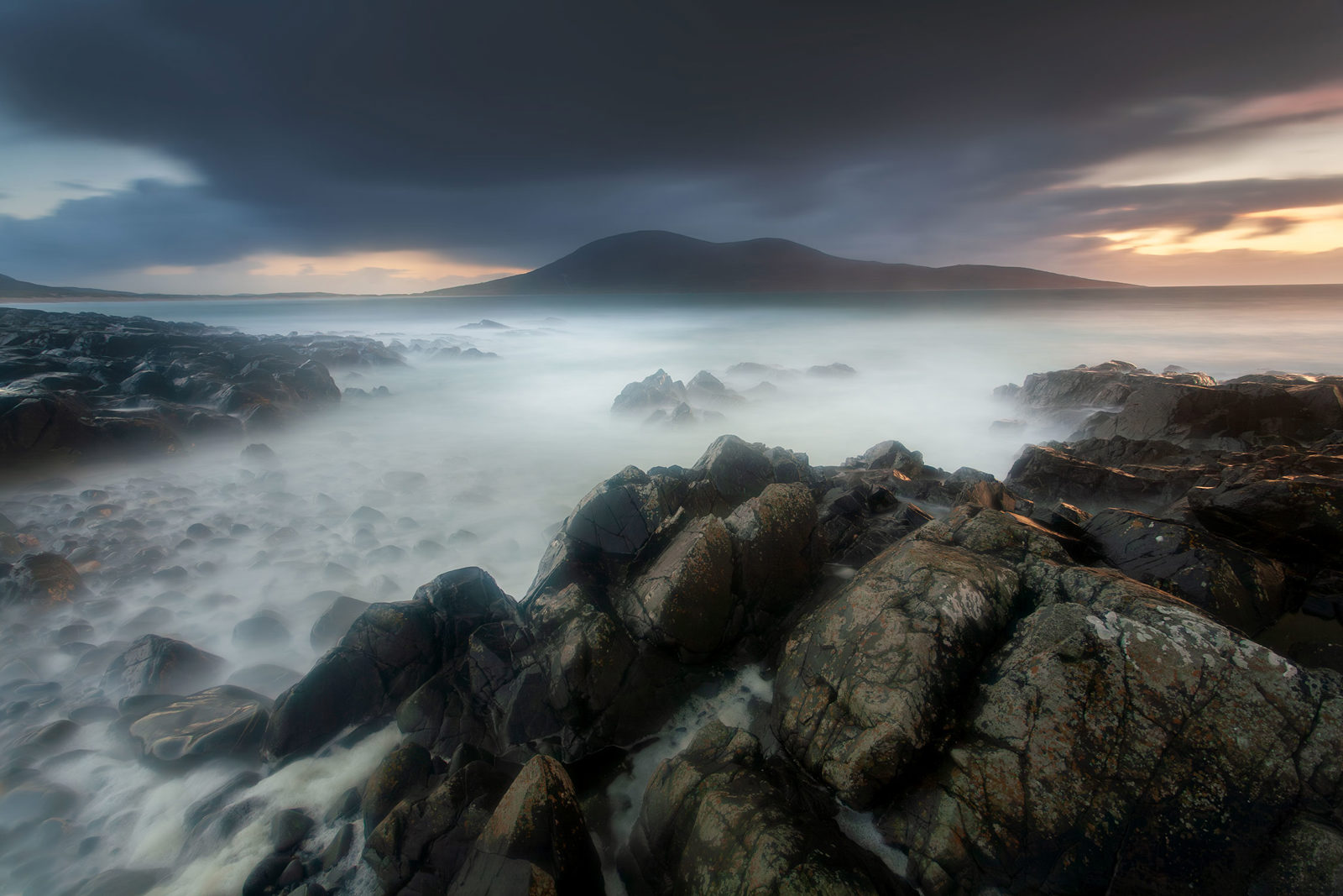 Misty seascape at twilight with rugged black rocks in the foreground. The sea appears smooth and ethereal due to long exposure, blending into the horizon. In the distance, dark hills are silhouetted against a dramatic, cloudy sky with a hint of light from the setting sun.