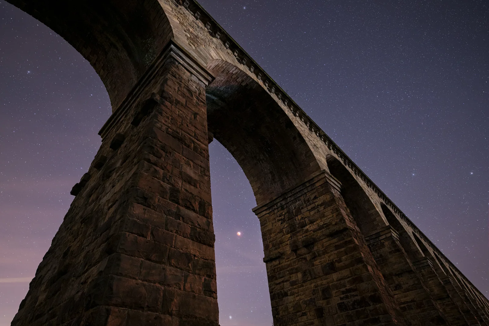 Historic stone railway viaduct arches silhouetted against a starry night sky with purple and blue hues.