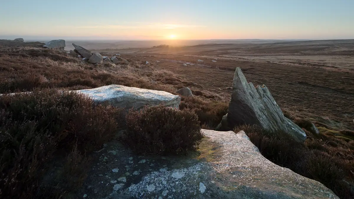 Sunset over a vast moorland with heather and rugged rock formations in the foreground. Sparse vegetation covers the rolling terrain, fading into distant hills under a clear sky with warm, glowing light from the setting sun.