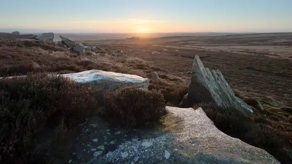 Sunset over a vast moorland with heather and rugged rock formations in the foreground. Sparse vegetation covers the rolling terrain, fading into distant hills under a clear sky with warm, glowing light from the setting sun.