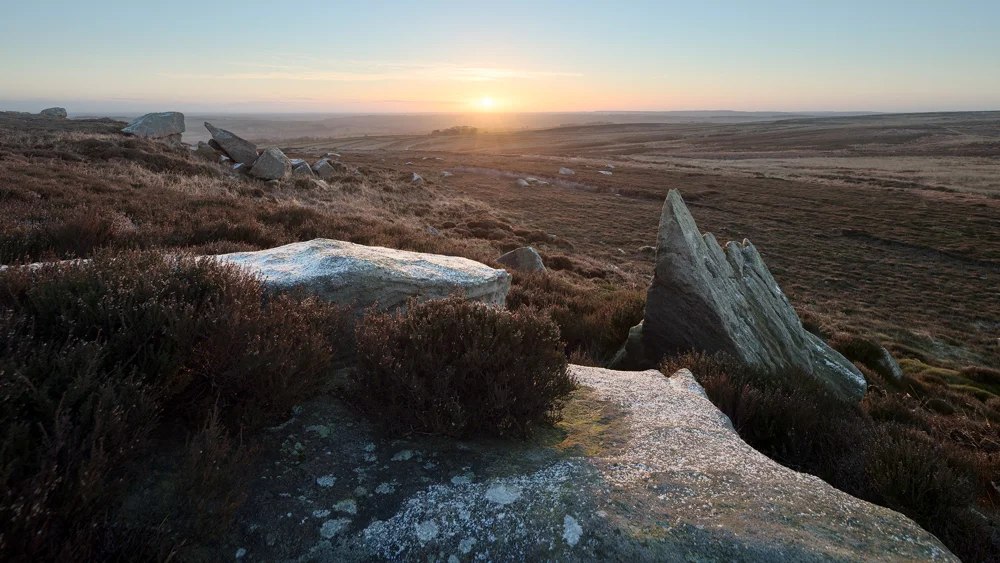 Lichen-covered gritstone boulders on open moorland at sunrise, with frost and golden light across heather