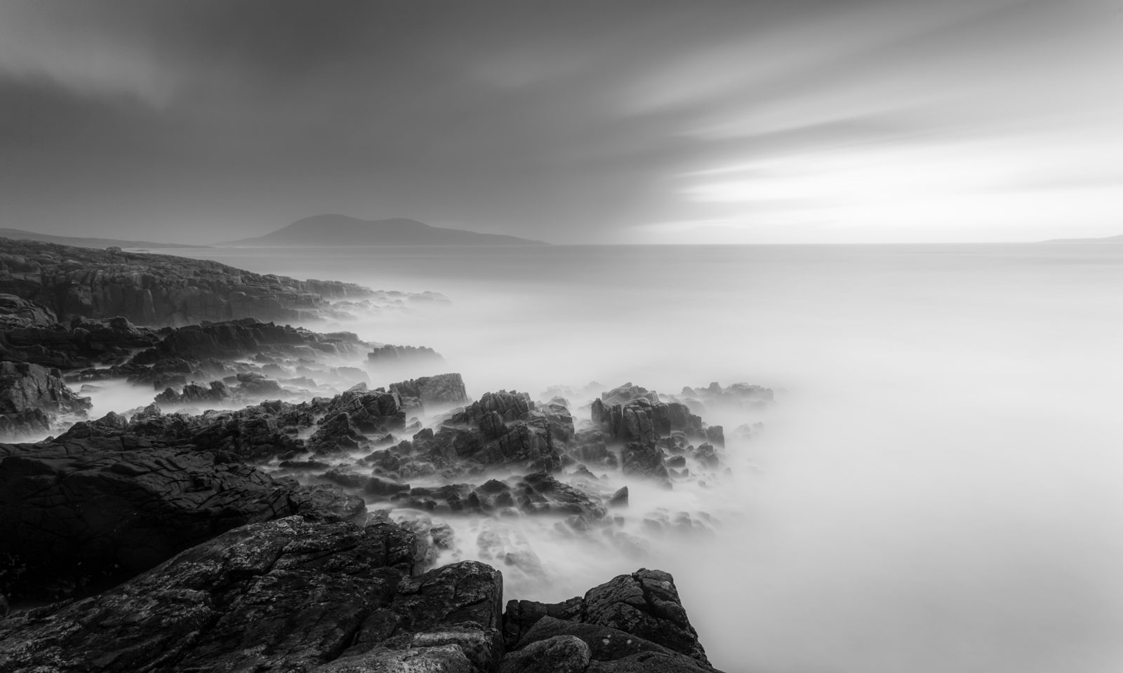 A monochrome seascape features an ethereal scene with mist swirling around jagged rocks leading to calm waters. In the distance, a mountain looms under a sky with streaked clouds, suggesting a time-lapse effect. The image conveys a serene yet dramatic atmosphere.
