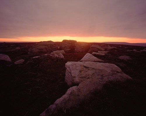 Moorland Landscapes: North Yorkshire moor at dusk. Large rocks and tufts of grass are silhouetted against the warm glow of a setting sun on the horizon, casting vivid hues of purple and orange across the sky. The scene conveys a tranquil, serene end to the day. a sunset over a snow covered mountain
