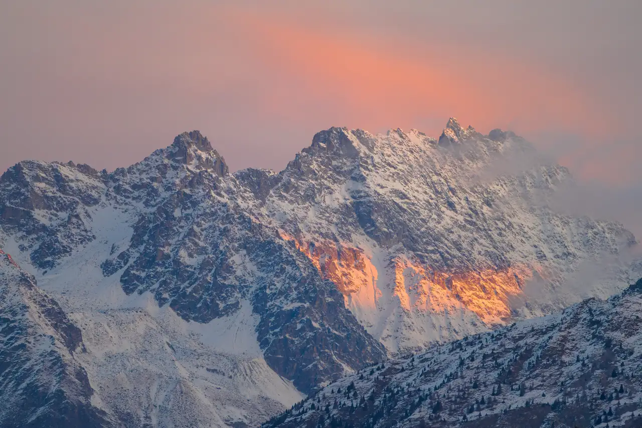 Snow-covered mountain peaks with a light dusting of snow, bathed in warm orange and pink hues from the setting sun. A pastel sky with soft clouds adds a gentle backdrop, enhancing the dramatic lighting on the rugged terrain. Sparse trees are visible on the lower slopes.