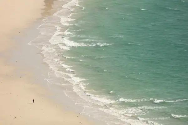 A lone person walks along a vast, empty beach with pale sand. Gentle waves from the turquoise sea lap against the shore, creating a white, foamy edge where the water meets the sand. The scene conveys a sense of solitude and tranquillity.