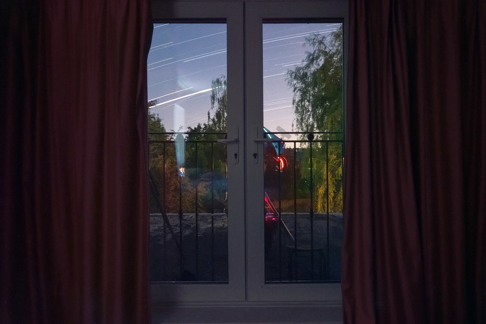 Star trails streaking across a twilight sky seen through French doors framed by deep red curtains