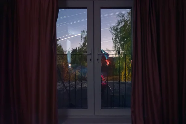 Star trails streaking across a twilight sky seen through French doors framed by deep red curtains