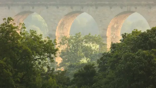 A stone viaduct with large archways, partially obscured by lush green trees in the foreground. Soft sunlight creates a warm glow on the viaduct, enhancing the stone texture, while a misty backdrop lends a serene atmosphere to the landscape.