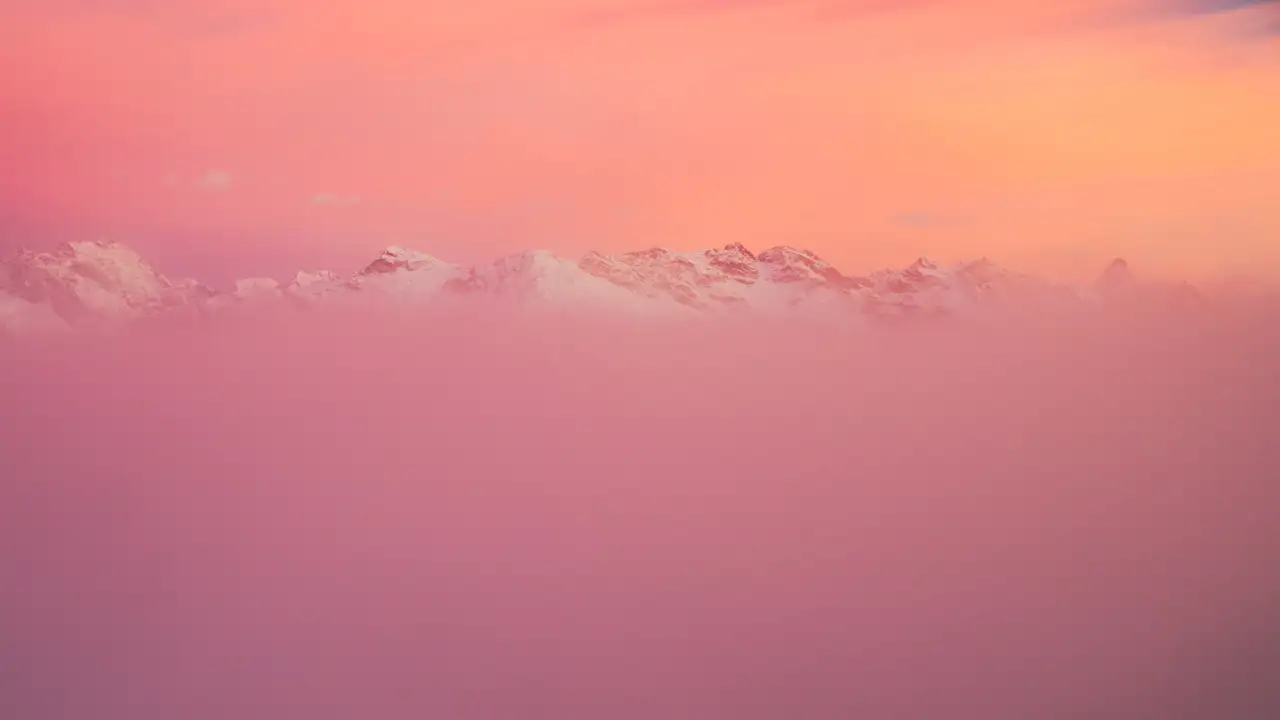 Snow-capped mountain peaks partially obscured by a layer of mist, with a vibrant pink and orange sky at sunset creating a serene, dreamy atmosphere.