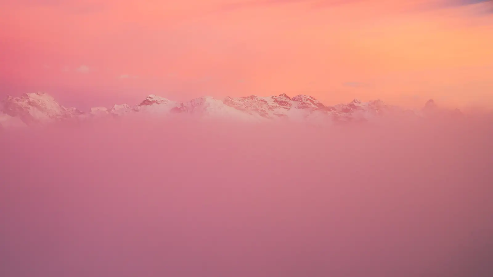 Snow-capped mountain peaks partially obscured by a layer of mist, with a vibrant pink and orange sky at sunset creating a serene, dreamy atmosphere.