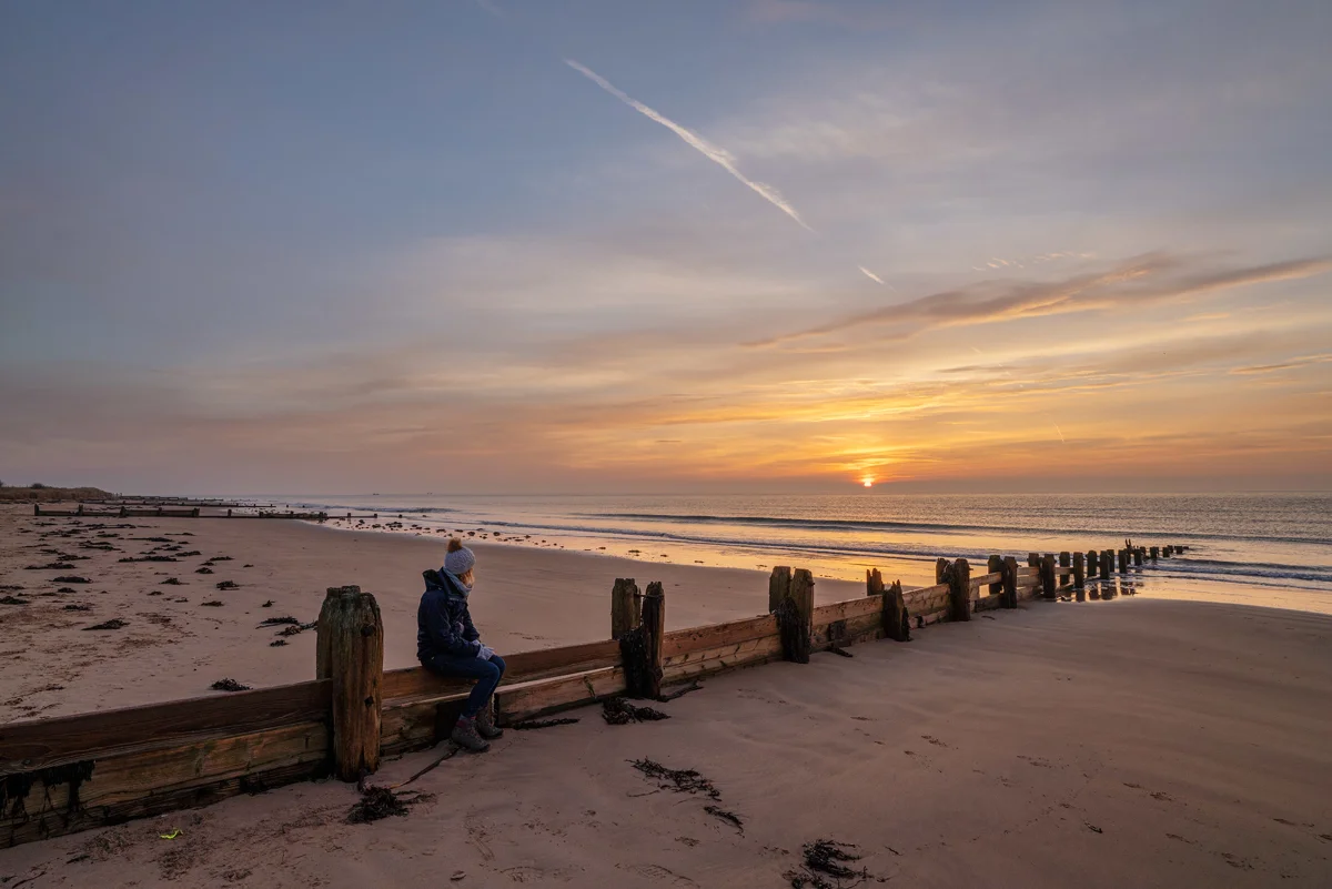 Person in bobble hat sitting on weathered wooden groyne on a sandy beach watching the sun rise over the sea