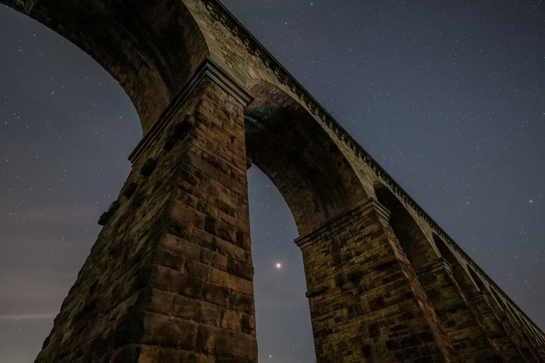 Stone railway viaduct arches lit from below against a star-filled night sky with a glowing planet