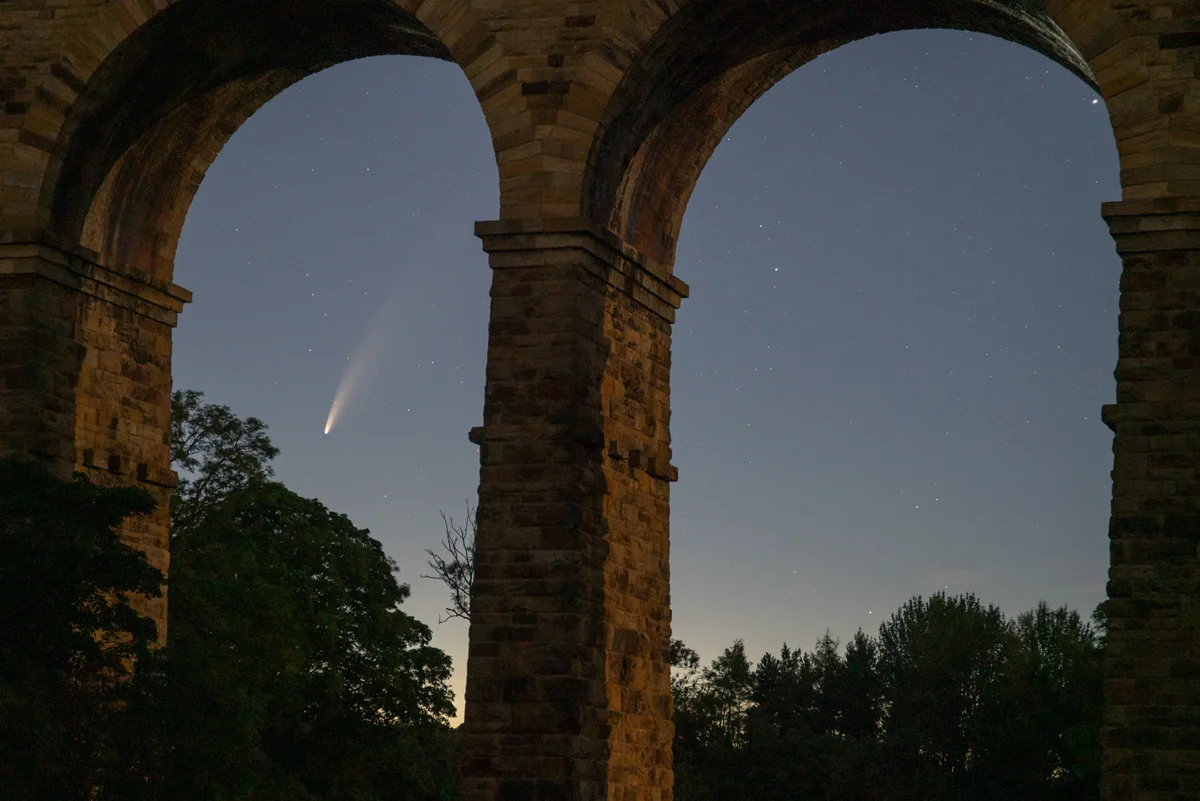 Comet Neowise glowing in a starry twilight sky between the stone arches of Crimple Viaduct, with trees silhouetted below.