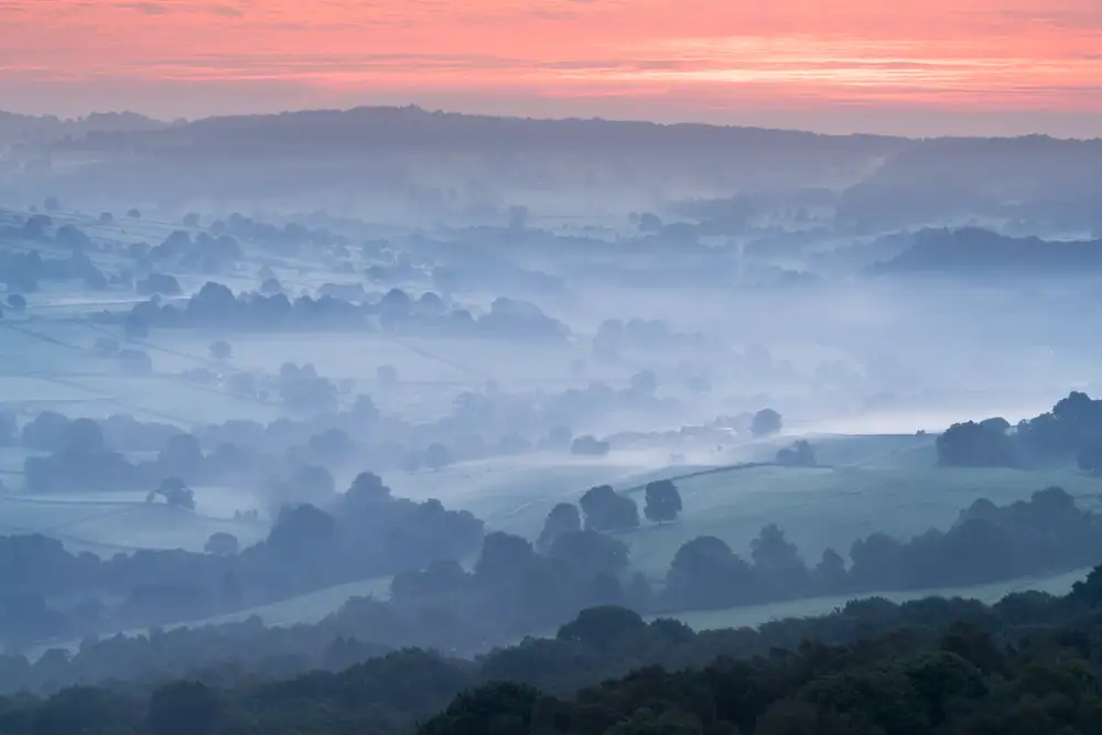 Rolling hills are covered in a soft morning mist, creating a serene and atmospheric scene. Patches of trees dot the landscape, and the mist blurs the boundaries between the fields. Above, the sky is painted with warm hues of pink and orange as the sun begins to rise, casting a gentle glow over the scene.