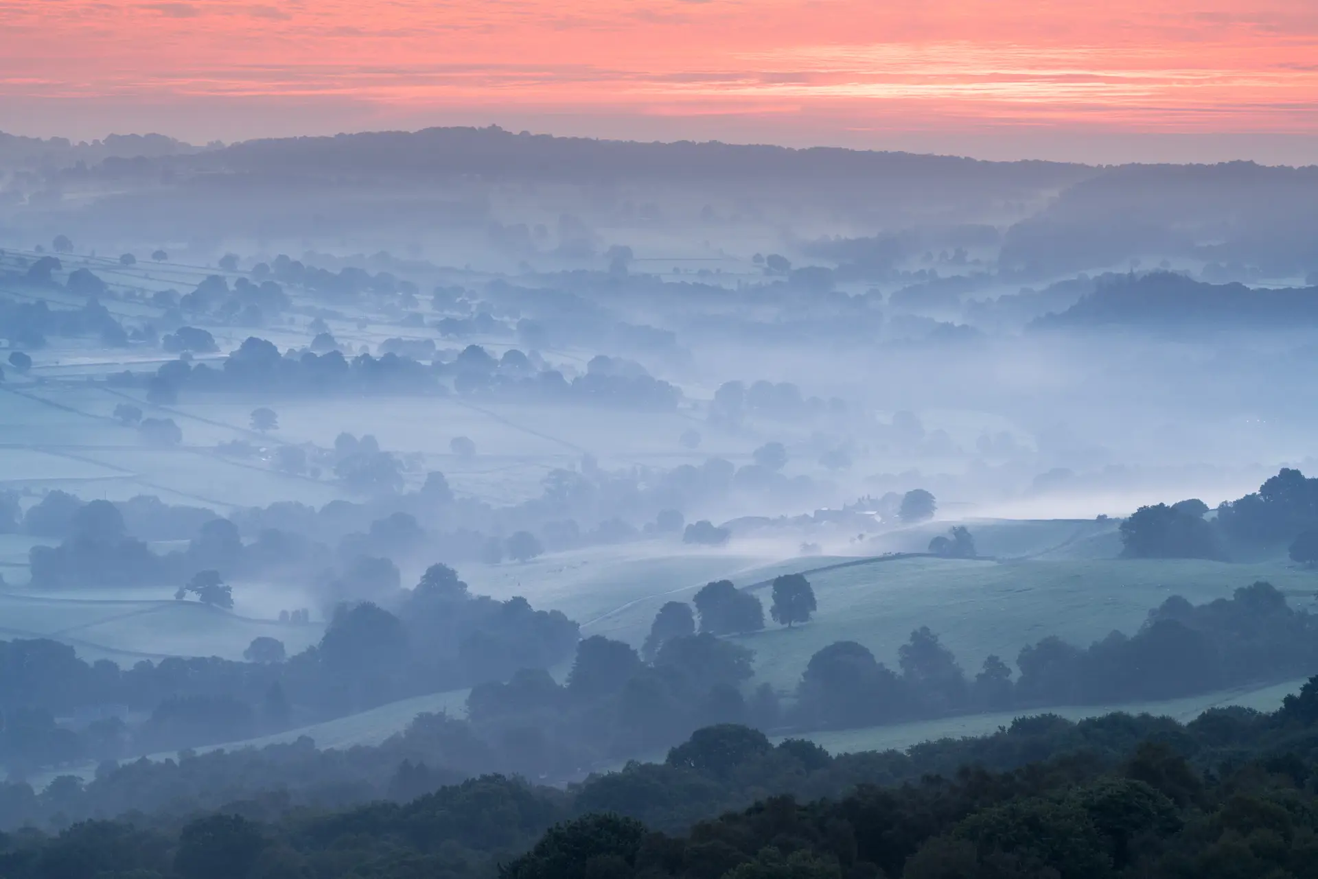 Rolling hills are covered in a soft morning mist, creating a serene and atmospheric scene. Patches of trees dot the landscape, and the mist blurs the boundaries between the fields. Above, the sky is painted with warm hues of pink and orange as the sun begins to rise, casting a gentle glow over the scene.