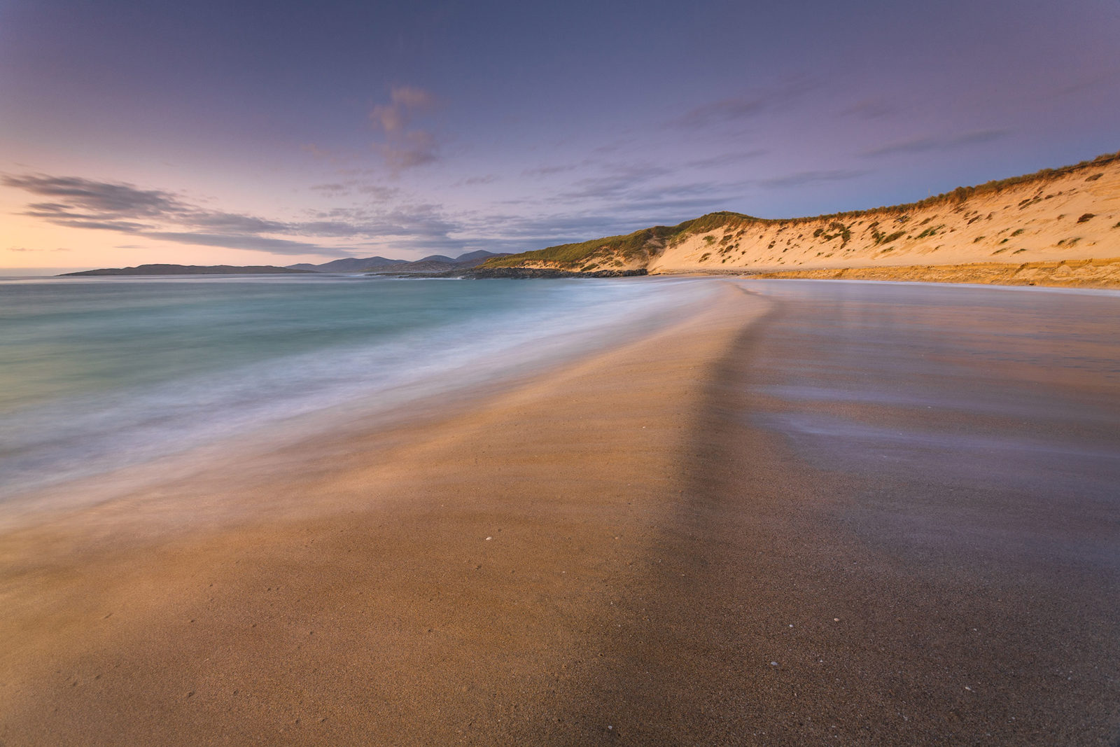 Tranquil beach scene at dusk with smooth, sandy shoreline curving into the distance on the right. The sea, coloured in soft blues and greens, gently laps the shore. Low, grassy sand dunes rise on the right, and a distant ridge of hills is silhouetted against the pastel sky, which transitions from pale orange to purple. Wispy clouds are scattered across the sky.