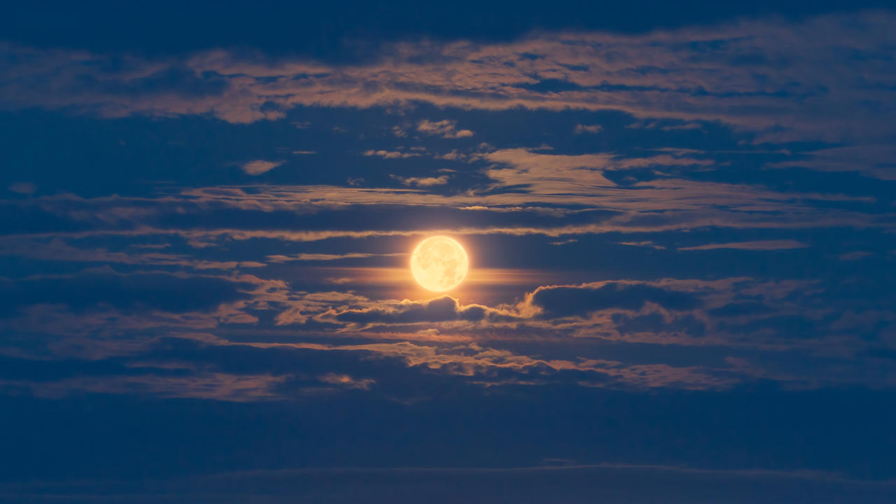 Bright full moon illuminating the night sky, partially obscured by scattered clouds, creating a soft, atmospheric glow. The clouds have subtle pink and blue hues, contributing to a serene and tranquil scene.