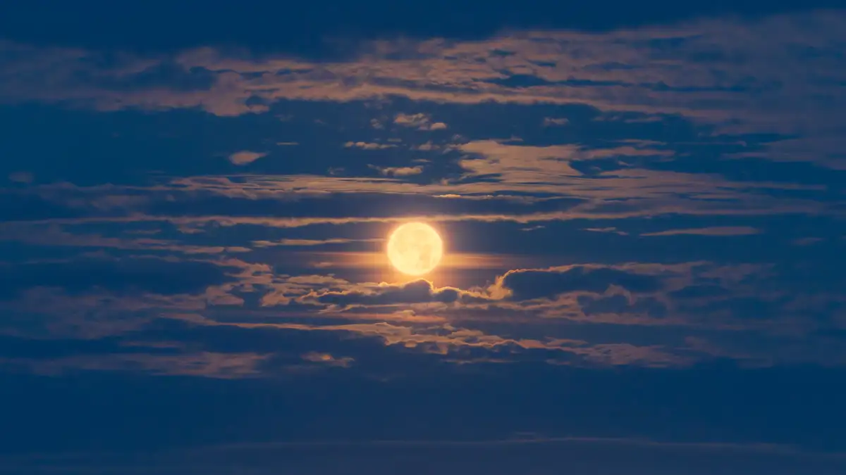 Bright full moon illuminating the night sky, partially obscured by scattered clouds, creating a soft, atmospheric glow. The clouds have subtle pink and blue hues, contributing to a serene and tranquil scene.