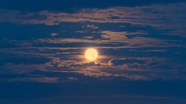 Bright full moon illuminating the night sky, partially obscured by scattered clouds, creating a soft, atmospheric glow. The clouds have subtle pink and blue hues, contributing to a serene and tranquil scene.